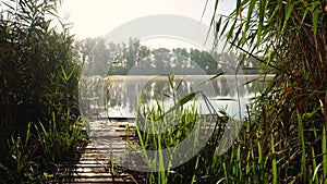 Sunrise on the riverbank. Landscape with wooden bridge among reeds and smoke on the water