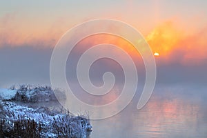 Sunrise time over the winter river. An orange fog spreads over the water and obscures the horizon. Reeds stand in the snow near