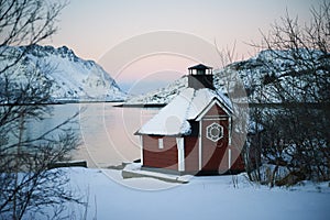 Sunrise, winter, unfrozen water, snow-capped mountains, red house and reflection of the pink sky,