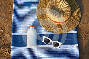 Sunscreen bottle with white sunglasses, straw on blue striped towel at beach signifies sun protection on sunny day by