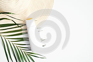 Sunscreen cream bottle on white background with female beach hat and tropical palm leaf. Flat lay, top view, copy space. Summer