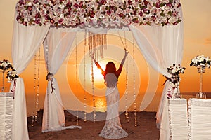 Sunset. bride silhouette. Wedding ceremony arch with flower arrangement and white curtain on cliff above sea, outdoor photo.