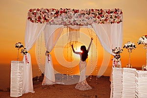 Sunset. bride silhouette. Wedding ceremony arch with flower arrangement and white curtain on cliff above sea, outdoor photo.