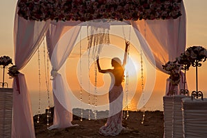 Sunset. bride silhouette. Wedding ceremony arch with flower arrangement and white curtain on cliff above sea, outdoor photo.