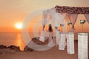 Sunset. bride silhouette. Wedding ceremony arch with flower arrangement and white curtain on cliff above sea, outdoor photo.