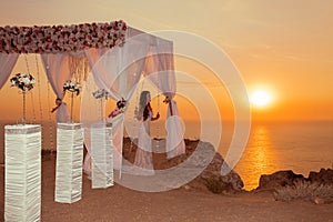 Sunset. bride silhouette. Wedding ceremony arch with flower arrangement and white curtain on cliff above sea, outdoor photo.