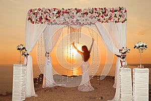 Sunset. bride silhouette. Wedding ceremony arch with flower arrangement and white curtain on cliff above sea, outdoor summer