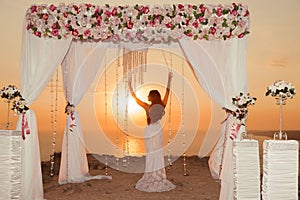 Sunset. bride silhouette. Wedding ceremony arch with flower arrangement, white curtain and crystals chandelier on cliff above sea