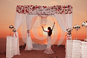 Sunset. Bride silhouette. Wedding ceremony arch with flower arrangement, white curtain and crystals chandelier on cliff above sea