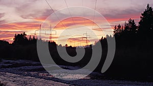 Sunset clouds over forest near river, silhouettes of electric power pylons and cables in distance