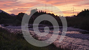 Sunset clouds over forest near river, silhouettes of electric power pylons and cables in distance