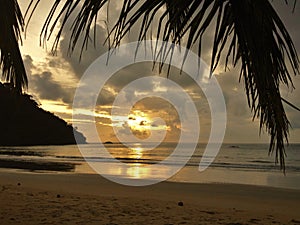 Sunset glow over a beautiful tropical beach and ocean water. Different colours of clouds and the silhouette of palm tree.