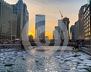 Sunset glow reflects off of windows of skyscrapers and ice on the surface of an icy Chicago River