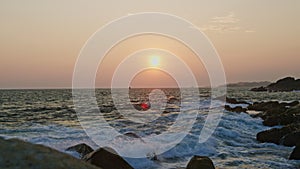 Sunset horizon over tumultuous ocean waves, rocks at foreground. Vibrant sky meets sea at dusk, reflecting on water