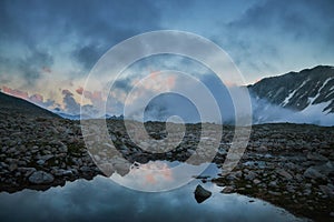 Sunset hues paint the sky as clouds roll over a calm mountain lake surrounded by rocky terrain