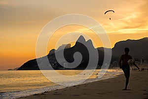 Sunset at Ipanema beach with Brazilian boy kicking a football and paraglider in sky with Dois IrmÃ£os Two Brothers mountains