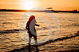 at sunset. kid girl walks along seashore, collects stones and throws into water