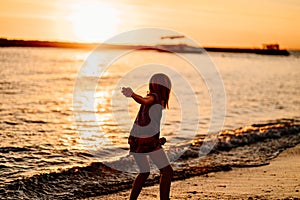 at sunset. kid girl walks along seashore, collects stones and throws into water