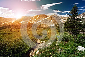 Sunset light shining behind a distant mountain range and a dirt hiking trail winding through an alpine meadow in Colorado