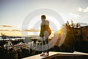 Sunset in Malaga, Spain. Young woman with backpack stands on a wall watching the horizon. Mediterrenean town and sea.