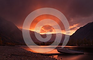 Sunset In The Mountains. Enchanting Autumn Mountain Landscape In Red Tones With Sunset Sky, River with Reflection And Lonely Boat.