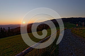 Sunset over country road and forested hills. Polish mountains. Rays of sun falling on a rural fence