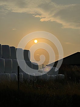 sunset over field of hay bales by barn with fence