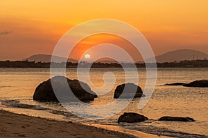 Sunset over land and ocean with reflections, silhouetted granite boulders and an orange glow