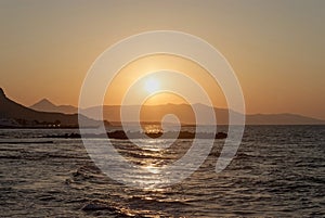 Sunset over the sea in Kokkini Hani, Crete, Greece. The sun disappears behind the mountain. Scenic seaside landscape in the