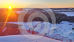 Sunset over a snow-covered ski and snowboard piste, equipped with chairlift and rope tow elevators, mesh fence and lampposts, surr