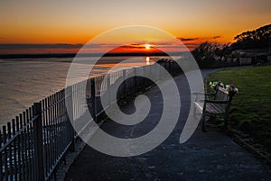 Sunset reflecting in the sea with the cliff top promenade and memorial bench in the foreground in soft focus