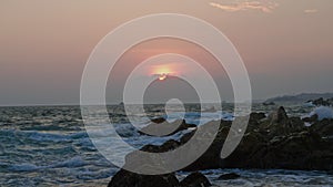 Sunset at sea with waves crashing on rocky shore. Light fades behind clouds above horizon. Coastal scene, tranquil