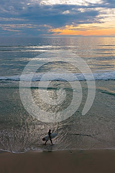 Sunset seascape, orange, blue, yellow sky, with aqua marine green sea, white waves rolling in, surfer at dusk walking in the surf