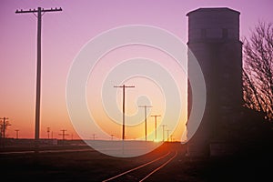 Sunset silhouette of grain silo beside train tracks, KS