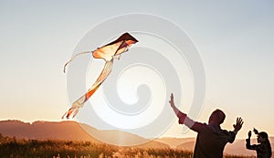 Sunset sun backlit silhouettes of a daughter with father while they trying to launch a colorful kite in the mountain fields. Warm