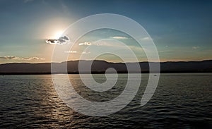 Sunset with sun partially obscured by clouds over lake and distant mountains.