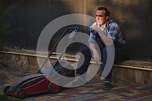 Sunset sun rays fall on face of a gardener man, landscaper with lawn mower, relaxing with cup of coffee in the backyard