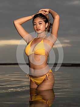 Sunset time on the beach. Beautiful Asian woman posing in yellow bikini. Tanned skin. Slim fit body. Water reflaction. Tropical