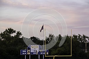 Sunset view of football end zone with goal posts, American flag flying and score board during evening