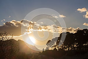 Sunset view with a sun hiding behind a mountain making tree silhouettes and brightening white clouds