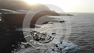 Sunset in volcanic lava formation with waves on wild beach and field of banana tree in greenhouses under the cliff.