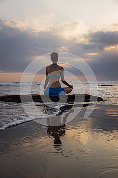 Sunset yoga. Caucasian woman sitting on the stone in Lotus pose. Padmasana. Hands in gyan mudra. Beach in Bali. View from back.