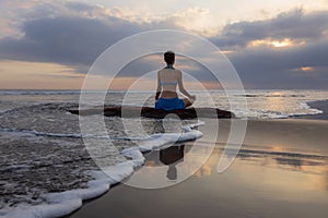 Sunset yoga. Caucasian woman sitting on the stone in Lotus pose. Padmasana. Hands in gyan mudra. Beach in Bali. View from back.