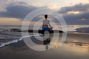 Sunset yoga. Caucasian woman sitting on the stone in Lotus pose. Padmasana. Hands in gyan mudra. Beach in Bali. View from back.