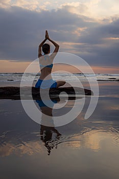 Sunset yoga. Caucasian woman sitting on the stone in Lotus pose. Padmasana. Hands in namaste mudra. Bali beach. View from back.