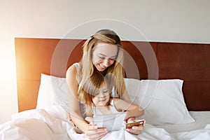 Sunshine portrait mother and little daughter doing online shopping at white bed with credit card, tablet