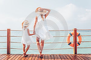 Suntanned woman and girl in white dresses enjoy sea view at the wooden pier
