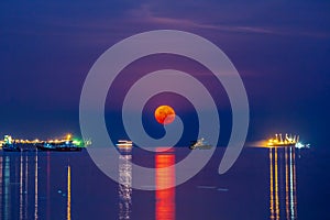 Super blood moon over Sriracha harbor with vessel cargo container ship on the sea water surface . night scene shot