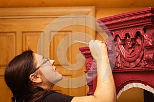 Super engaged lady painting cupboard made of wood with splendid carved ornaments in red color with brush looking at