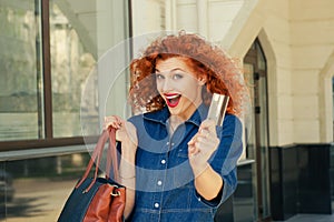 Super excited woman holding showing credit card bag near clothing department store shopping mall outdoor. Curly red head happy